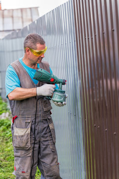 Positive man in overalls and protective glasses with spray gun in hand. Painting of metal fence