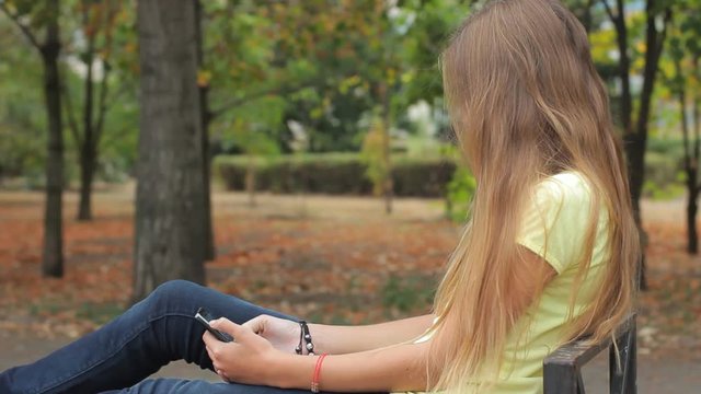 Sad teen girl texting using smart phone on park bench