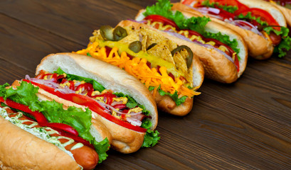 A lot of big delicious hot dogs with sauce and vegetables on wooden background. Their assorted hot...