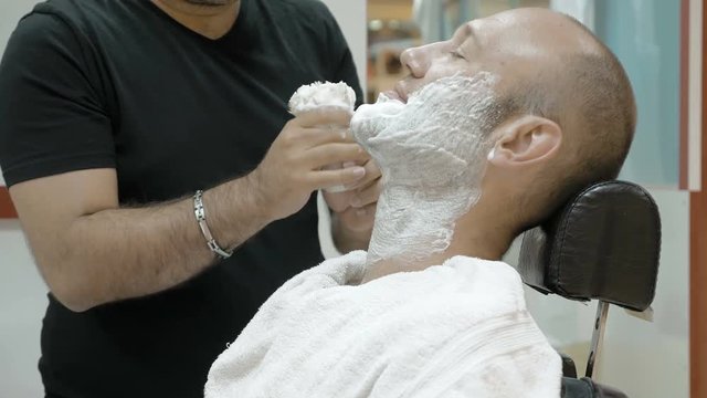 A man is sitting at a barber's shop to get groomed. The barber is lathering on the shaving cream on his client with a soft blending brush.