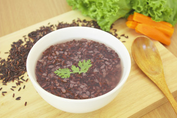 gruel (dark violet rice) in white plastic bowl  with carrot and lettuce and wooden spoon on wooden cutting board 