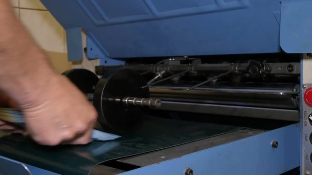Worker picks up last sheets of paper that came out from under roller, and turns off conveyor of folding machine. Related to printing, typography, work at printing house. Closeup shot. 4K Ultra HD.
