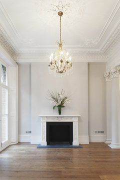 Grand drawing room, double french doors framed by pillars leading to balcony with a marble fireplace, chandelier and ornate plaster moulding.