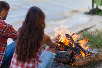 friendship, happiness, summer vacation, holidays and people concept - close up of couple sitting near fire on beach