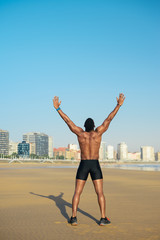 Fototapeta na wymiar Sport motivation and freedom concept. Back view of strong fit black man raising arms towards the city at the beach for celebrating running workout success.