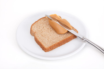 Peanut Butter Spread on Wheat Bread – A slice of wheat bread with knife spreading peanut butter. One a white plate, on a white background.