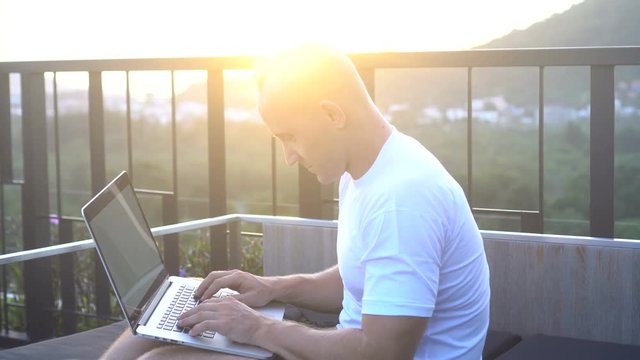 Outdoors portrait of handsome young man in sunglasses working on a laptop computer while sitting on a rooftop over beautiful sunrise. Freelance and technology
