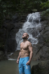 Handsome man with beard wearing blue shorts standing and looking up near waterfall. Male tourist enjoying by a water fall in forest