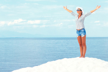 Young woman standing on white beach with her arms outstretched over horizon. She is looking towards the sun.