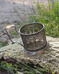 patterned silver stand for glasses,slightly covered with patina, stands on a piece of bark from the tree, near the grass, Sunny day,with a little sunlight