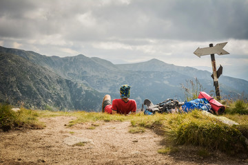 Direction indicator and tourist sitting in the grass looking. Mountain peaks in the background