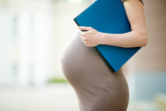 Active pregnancy concept. Close-up of belly of happy young business pregnant woman standing on the street with document folder. Future mom working on her late pregnancy period