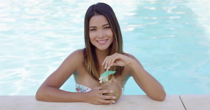 Pretty young woman relaxing in a resort swimming pool cooling off in the water while enjoying a tropical coconut cocktail and smiling at the camera
