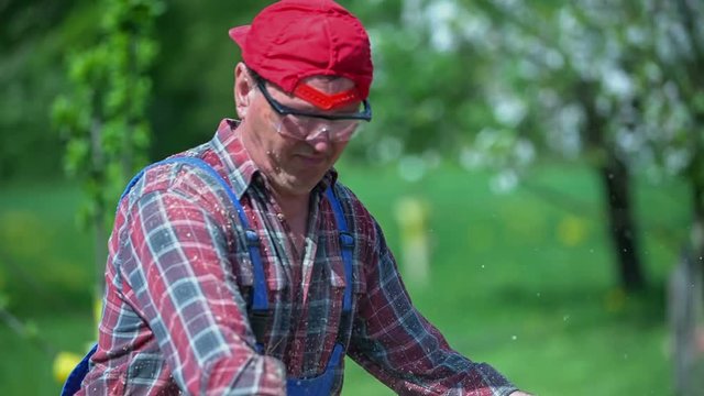 A joiner is cutting wood with the help of a circular saw. It is a nice sunny day and he is wearing a colourful shirt and a cap which is turned backwards.
