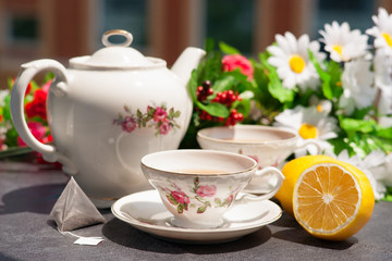 Чай в чайном сервизе, чайный пакетик и лимон на фоне цветов и окон здания на балконе.