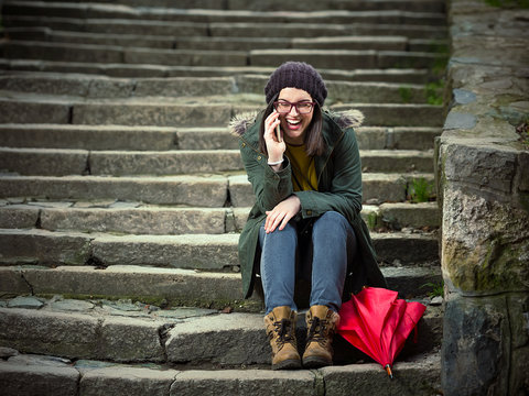 Happy young hipster woman is sitting on the outdoors stairs, smiling and talking on the mobile phone.