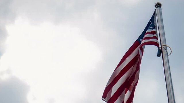 United States American flag flying from a flagpole in a wind showing the stars and stripes against a cloudy blue sky in a patriotic image. View from low angle