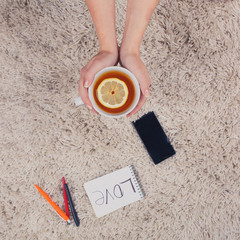 A woman holds hands a cup of tea with lemon, phone, pen and notebook lying on the carpet, top view