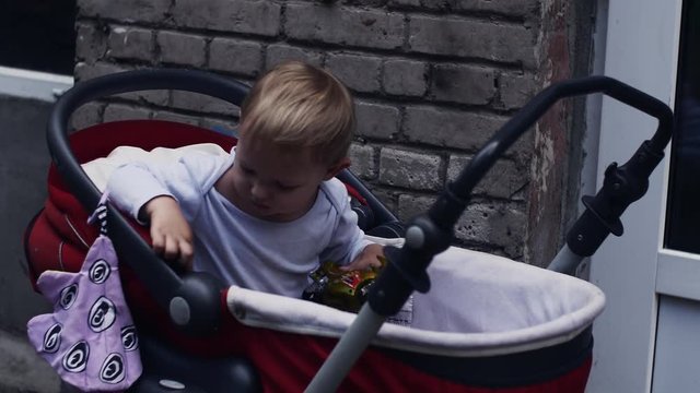 Little boy play with toy in red baby carriage on street. Summer walking. Child.