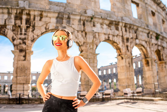 Young sports woman with headphones and smartwatch resting after the training near the ancient coliseum in Pula city