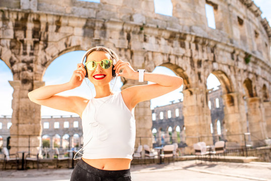 Young smiling sports woman listening to the music with headphones standing near the ancient amphitheatre in Pula city.