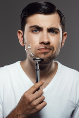 Displeased young handsome man shaving over grey background.