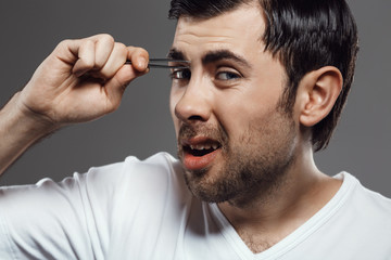 Young handsome man tweezing eyebrows over grey background.