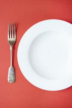 An empty white dinner plate and fork on a red background
