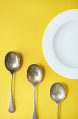 An empty white bowl and silver spoons arranged on a yellow background