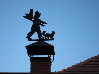 Silhouette of a chimney sweep with cats and stairs to the flue pipe of red brick in front of the blue sky