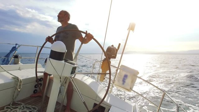 Mature adult skipper driving a huge sailboat on sunrise / sunset at the ocean. The sun is shining bright from behind