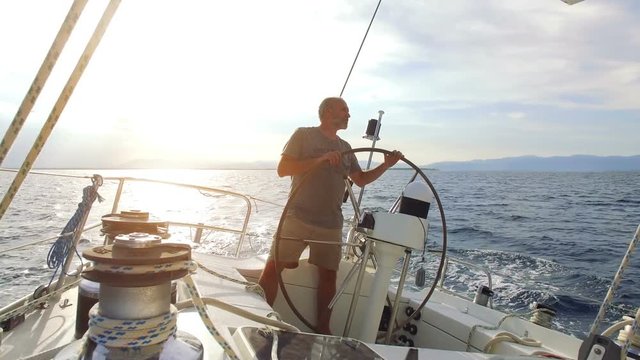 Mature adult skipper driving a huge sailboat on sunrise / sunset at the ocean. The sun is shining bright from behind