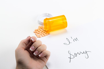 Pills Spilling out of Bottle near Womans hand near sorry note, implication suicide overdose, posed studio shot