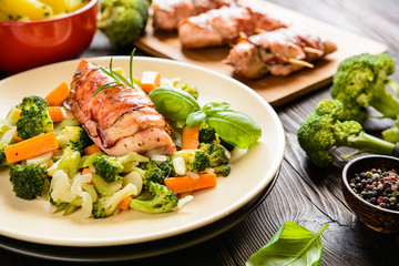 Baked chicken breast wrapped in bacon slices with boiled potatoes and steamed broccoli, carrot, garlic, onion