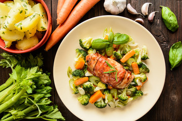 Baked chicken breast wrapped in bacon slices with boiled potatoes and steamed broccoli, carrot, garlic, onion