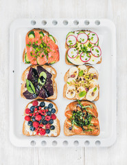 Sweet and savory breakfast toasts assortment. Sandwiches with fruit, vegetables, eggs and smoked salmon on white baking tray over white painted wooden background, top view © sonyakamoz