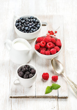 Fresh rasberries, blackberries and bilberries and milk in pitcher on white painted wooden background, top view, selective focus, vertical composition