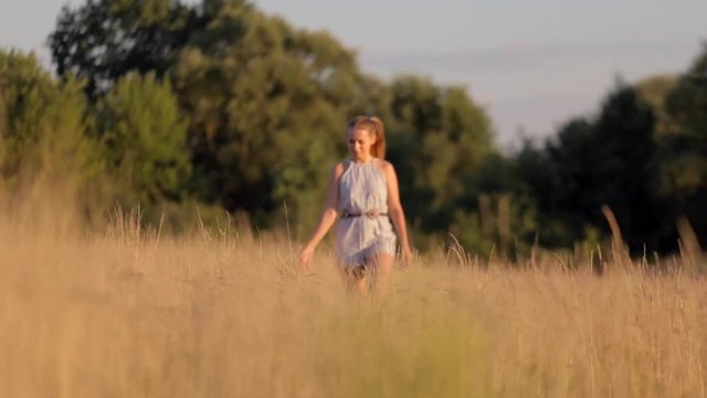 Beautiful young girl walking on the field with tall grass in the rays of the setting sun