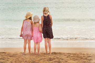 3 sisters stand on beachfront facing the ocean.