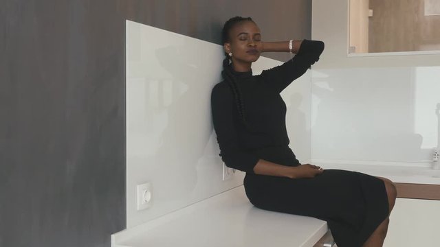 Serious African-American woman dressed in black sitting on the table leaned against the wall