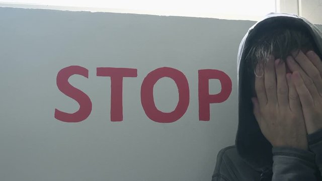 Hopeless drug addict standing next to stop sign on the wall, hand covering eyes and face in despair, man having drug addiction related problems.