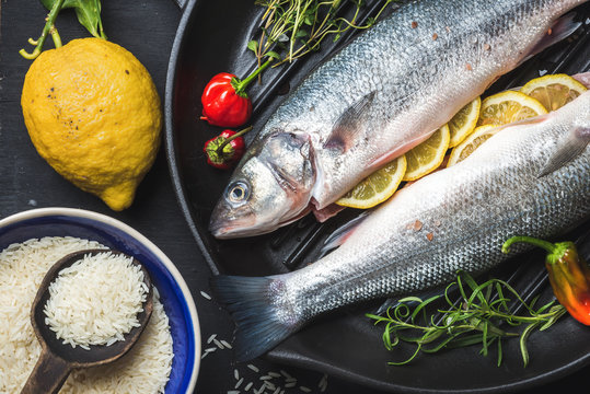 Raw uncooked seabass fish with rice, lemon, herbs and spices on black grilling iron pan over dark background, top view, horizontal composition