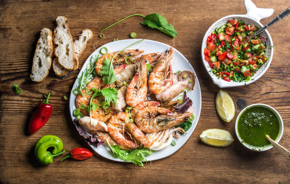 Plate of tiger prawns and pieces of octopus with fresh leek, green salad, peppers, lemon, bread and pesto sauce over wooden background, top view, horizontal composition