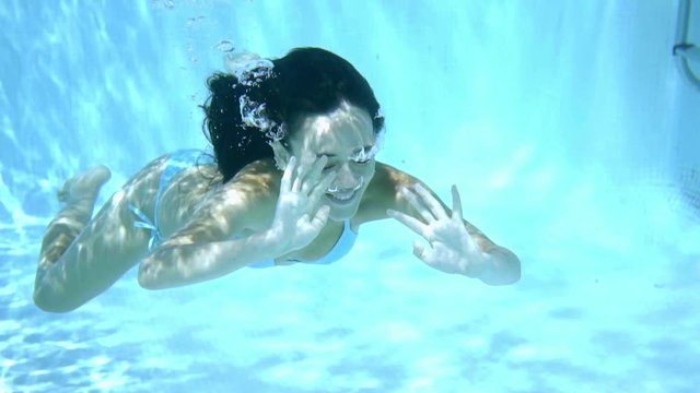Young woman swimming underwater in a swimming pool with reflections of the sunlight with closed eyes and arms outstretched in a close up view