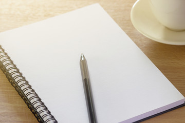Blank notebook on the wooden desk background with a pen on top and a coffee cup the right side.