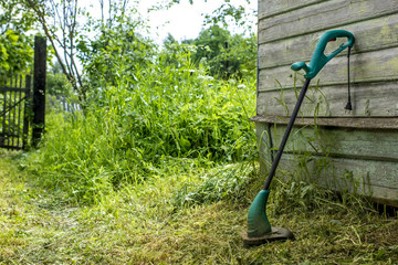 electric grass trimmer stands in the garden near the house, simply supported on the wall