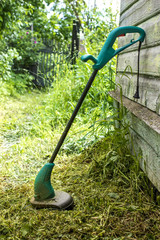 electric grass trimmer stands in the garden near the house, simply supported on the wall