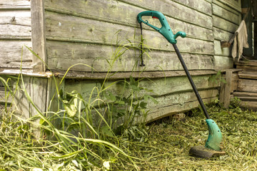 electric grass trimmer stands in the garden near the house, simply supported on the wall