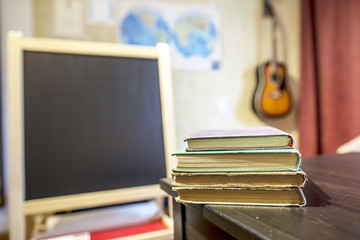 Books lying on a table in a classroom at a private school with a black board, card and guitar on the wall