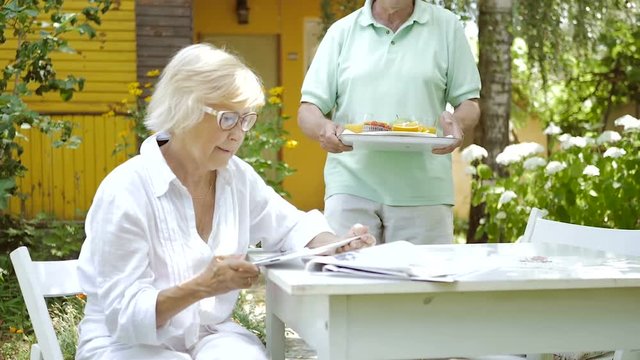 An old man brings a tray with lunch in the garden.